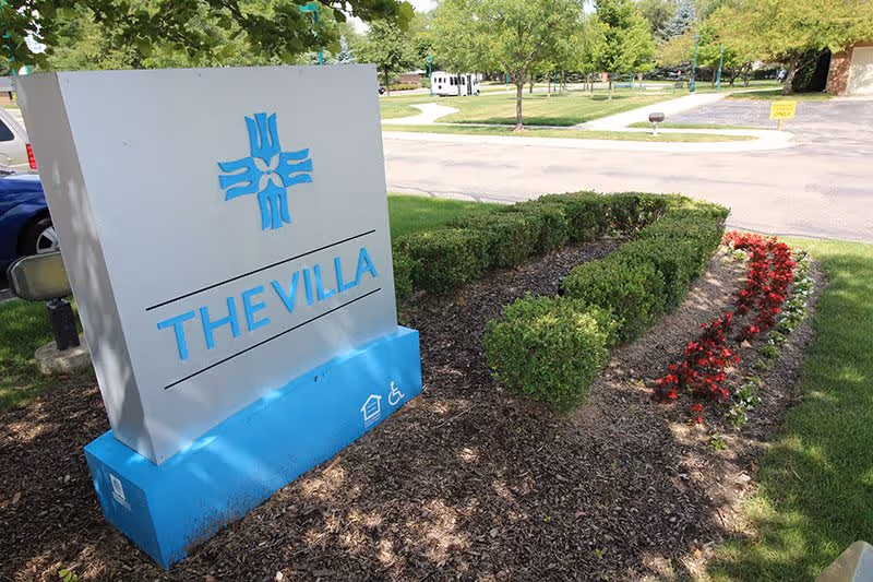 Outdoor view of a large sign for The Villa, situated on a landscaped area with trimmed bushes and red flowers, with a parking lot and trees in the background.