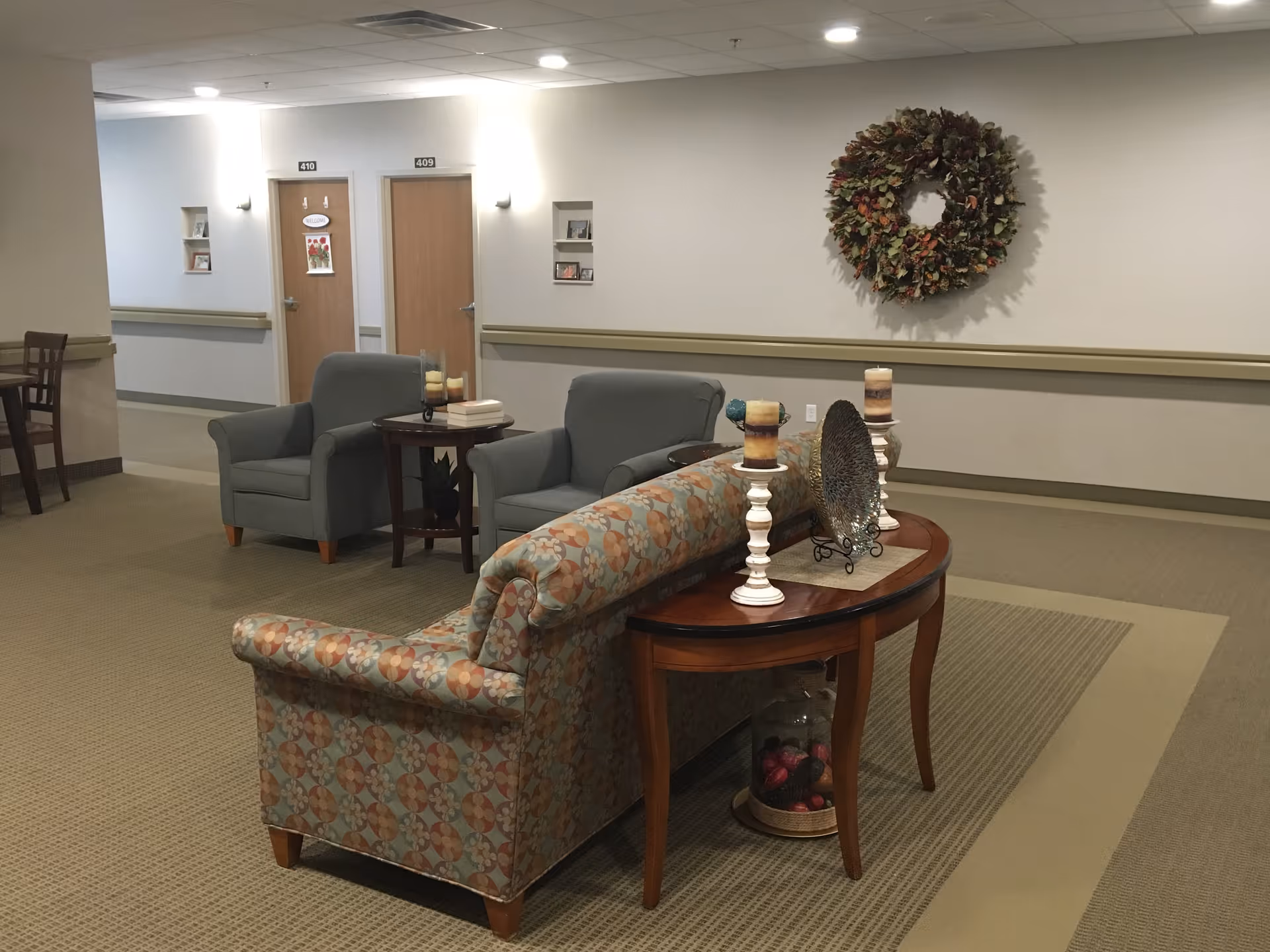 A sitting area in a hallway of an assisted living facility with two gray armchairs, a patterned sofa, a wooden side table with candles and decorative items, and a large wreath hanging on the wall. Two doors labeled 409 and 410 are visible in the background.