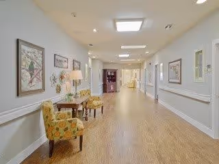 A well-lit hallway in a senior living facility with wooden flooring and light-colored walls. The hallway features decorative framed artwork and wall-mounted lamps. Two patterned armchairs and a small table with a lamp are placed along the left wall. The corridor extends into the distance with doors and additional seating visible.
