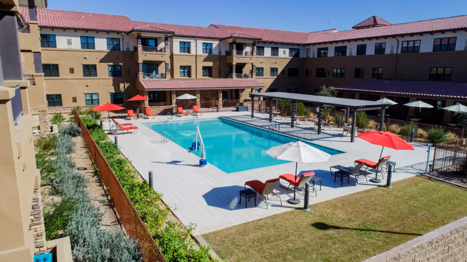 Sunny courtyard with a rectangular swimming pool, red and white lounge chairs and umbrellas, and a surrounding three-story residential building.