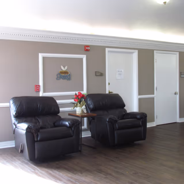 Two black leather recliner chairs with a small wooden side table between them holding a vase of red flowers, set against a beige wall with white trim and two closed white doors in the background.