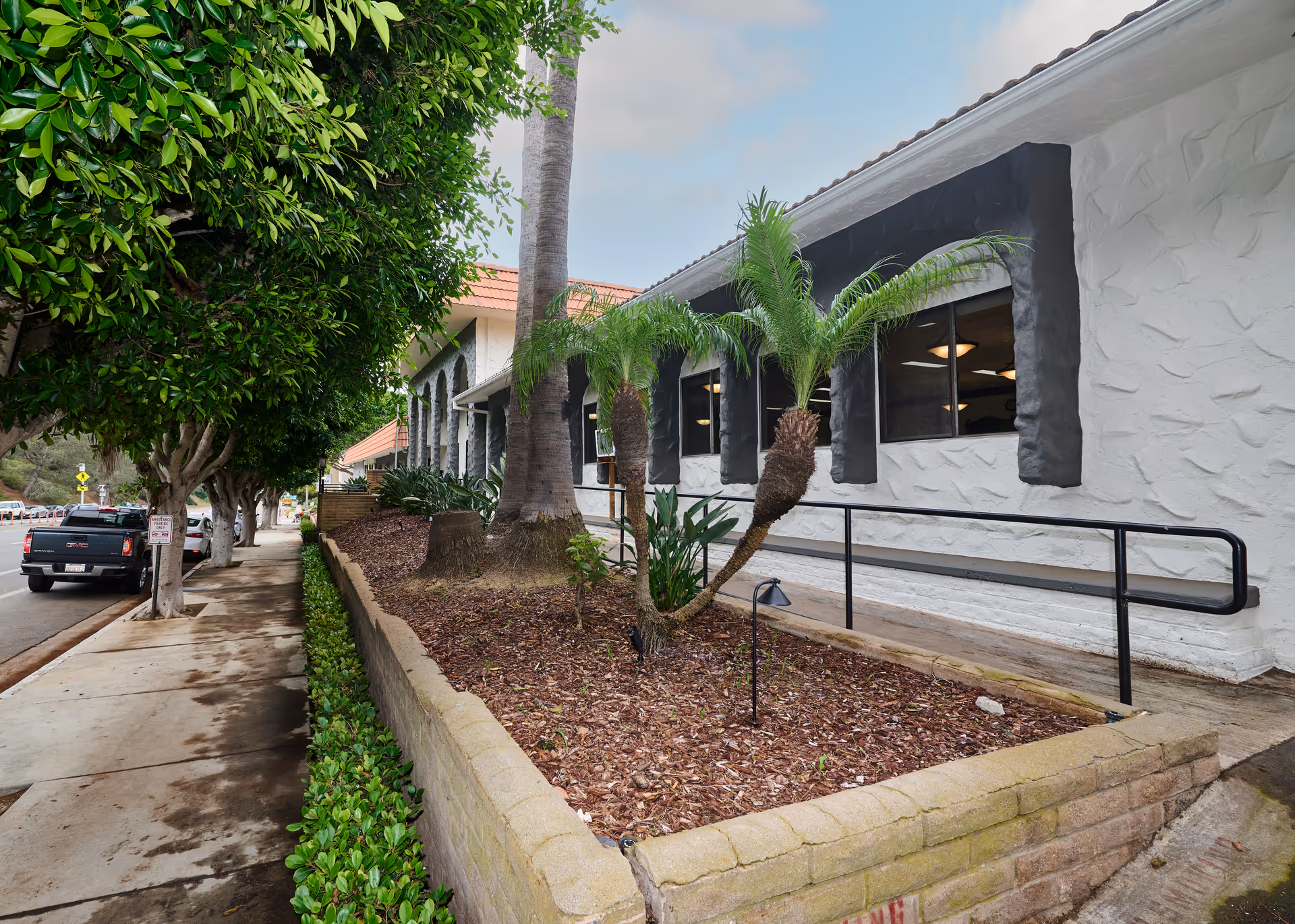 Front exterior of a white stucco building with black-trimmed windows, palm trees, a wheelchair ramp and a sidewalk with parked vehicles.