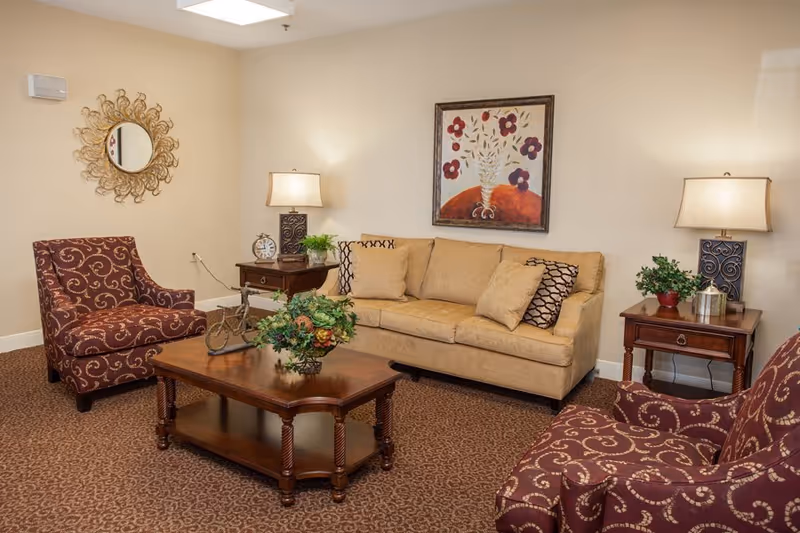 A cozy living room with a beige sofa adorned with patterned cushions, two maroon armchairs with gold swirl designs, a wooden coffee table with a floral centerpiece, two wooden side tables each with a lamp and decorative items, a round ornate mirror on the wall, and a framed floral painting above the sofa.
