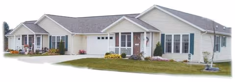 Single-story residential building with beige siding and multiple windows, featuring a small porch with white railings and a garage door, surrounded by a well-maintained lawn and some flower beds.