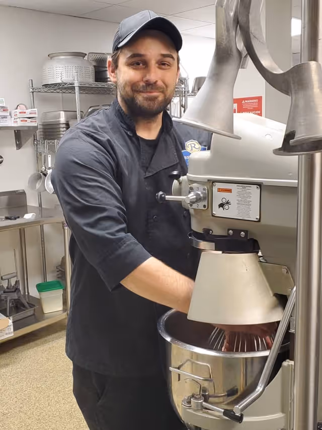 A man wearing a black chef's coat and a black cap is standing in a kitchen, operating a large industrial mixer. Shelves with kitchen utensils and containers are visible in the background.