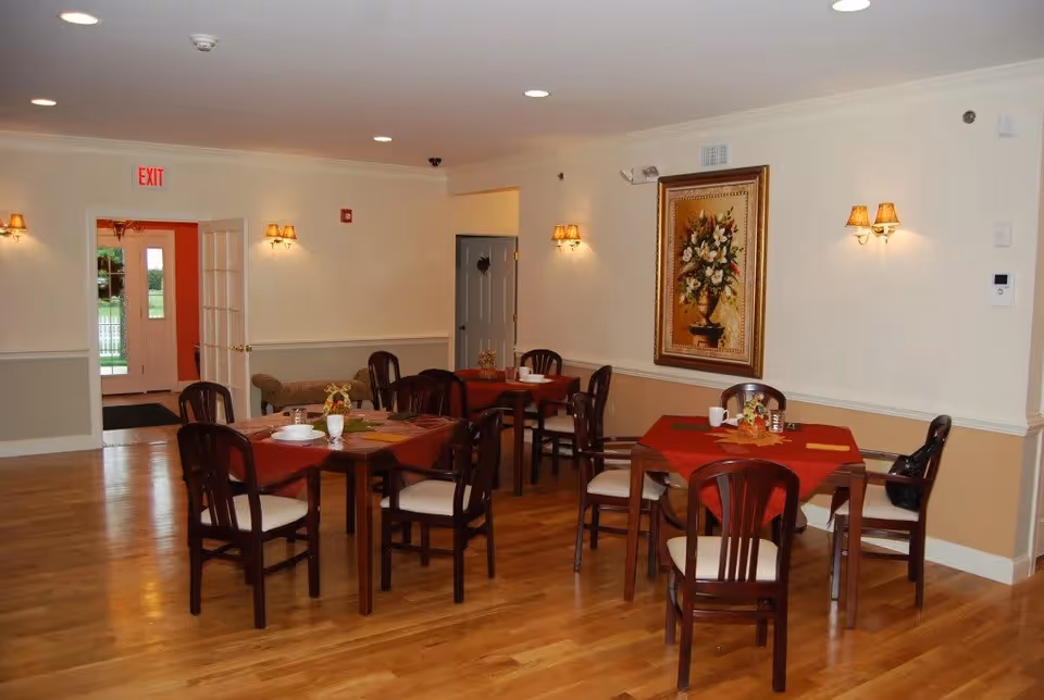 Interior dining room with wooden floors, several tables covered with red tablecloths, each surrounded by wooden chairs with white cushions. The walls are painted beige and white, adorned with wall sconces and a large framed floral painting. An exit door with a red exit sign is visible in the background.