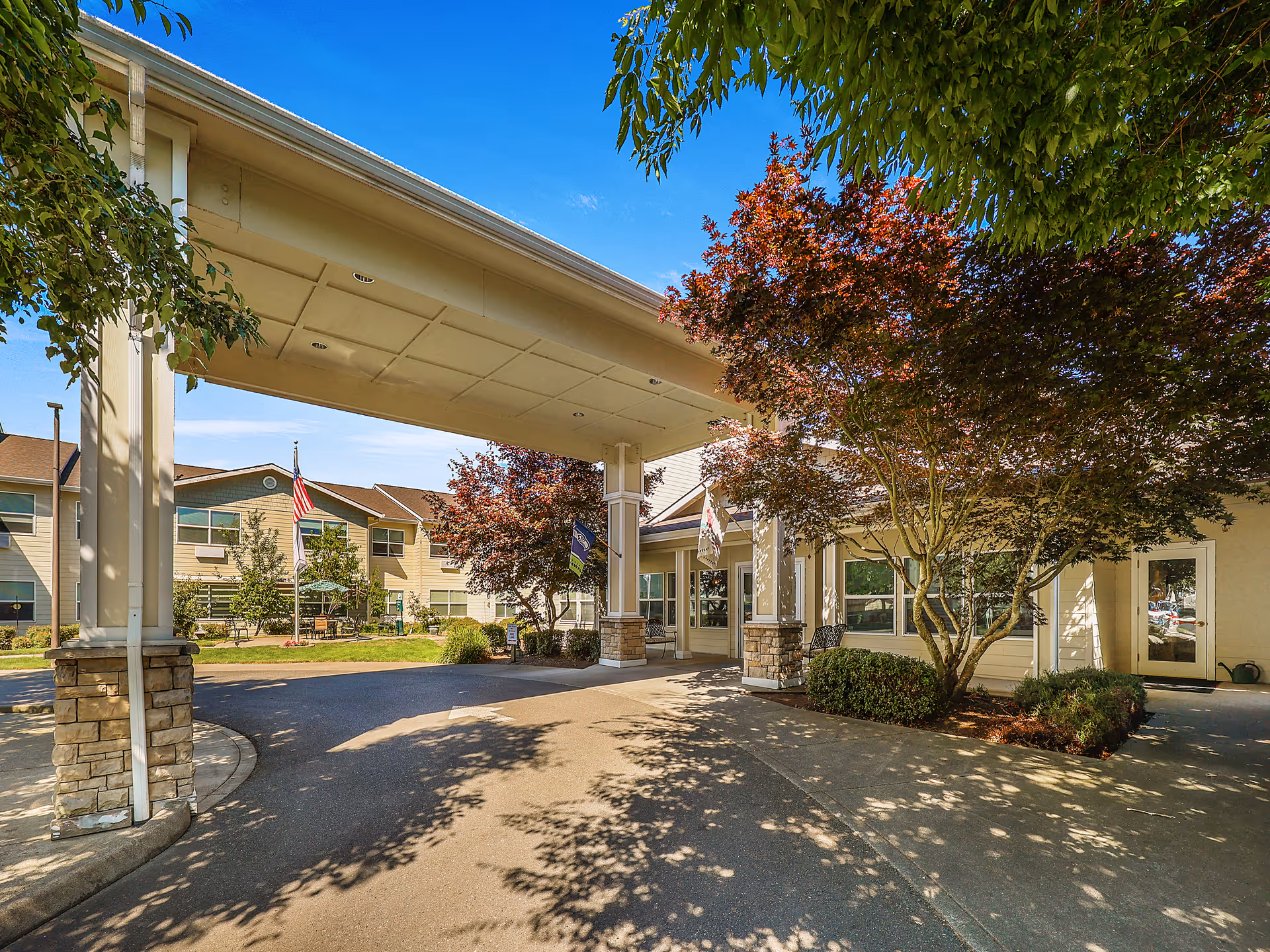 Entrance of Prestige Senior Living Auburn Meadows featuring a covered driveway with stone pillars, surrounded by trees with red and green leaves, and a two-story building in the background under a clear blue sky.