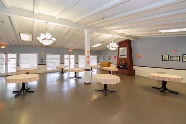 A spacious common area with several round tables covered with light pink tablecloths, a brick fireplace with a framed picture above it, a piano in the corner, and multiple windows with blinds letting in natural light. The ceiling has exposed beams and two chandeliers hanging down. The walls are decorated with colorful flower cutouts.