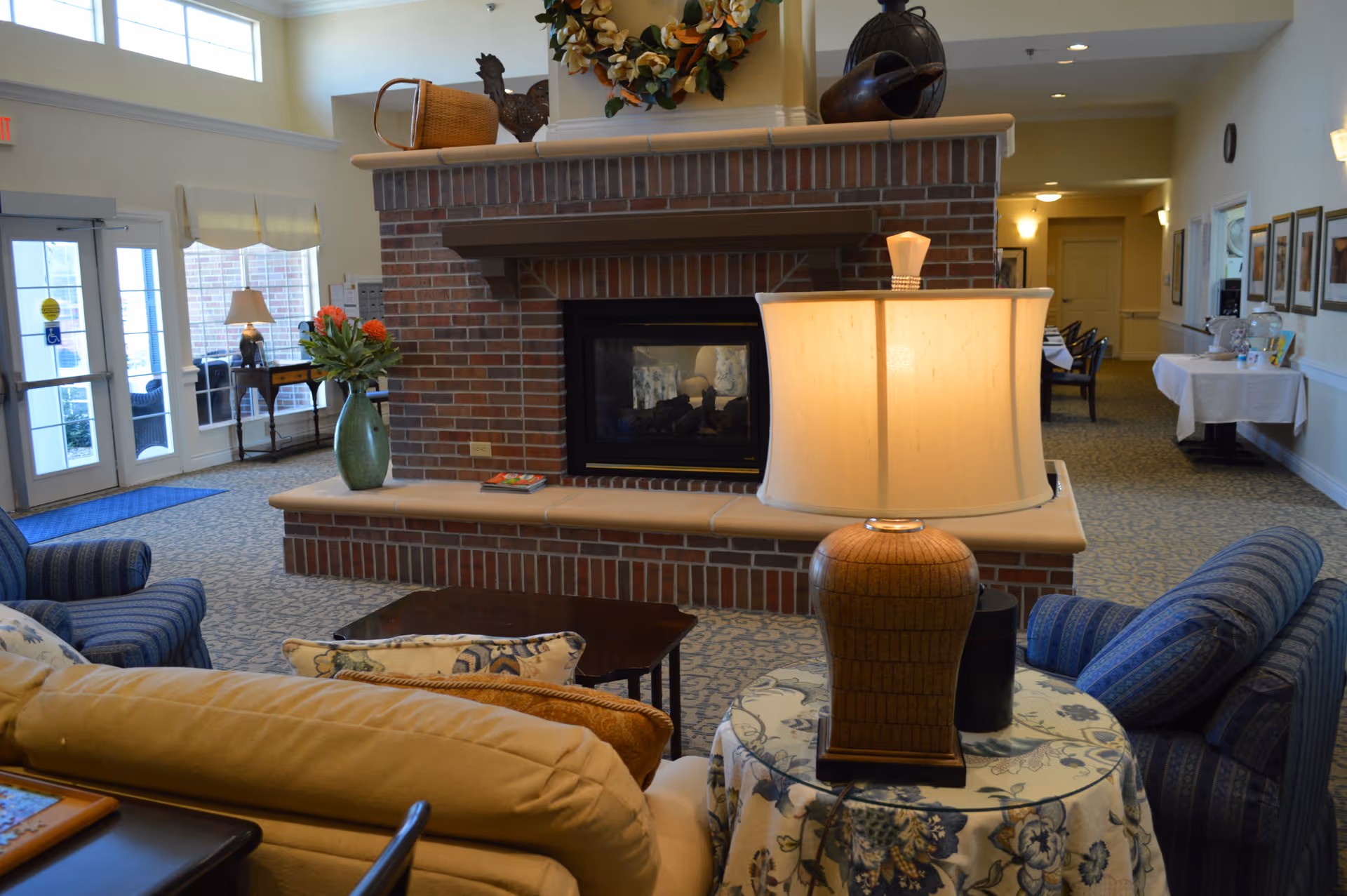 Communal living area with a central brick fireplace, upholstered chairs, a side table with a lamp, and floral decor.