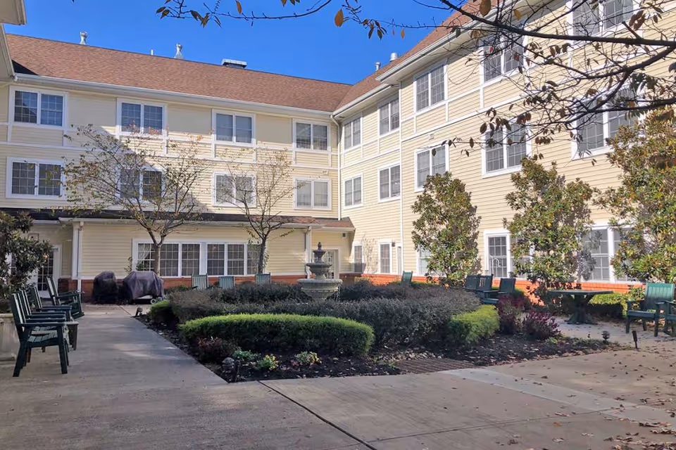 Outdoor courtyard area of a senior living facility with a central fountain surrounded by trimmed bushes and small trees. The courtyard is enclosed by a three-story beige building with many windows. Green chairs and benches are placed along the concrete pathways around the garden area under a clear blue sky.