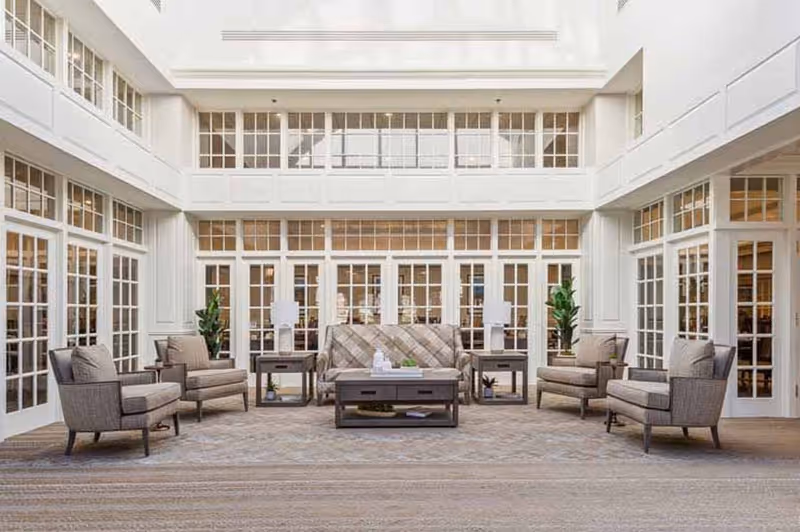 A spacious and bright living room area with large windows and glass doors surrounding the space. The room features a central sofa with a patterned design, flanked by four cushioned armchairs arranged symmetrically. There are two side tables with lamps and decorative plants placed near the walls. The floor is covered with a large area rug, and the overall color scheme is neutral and calming.