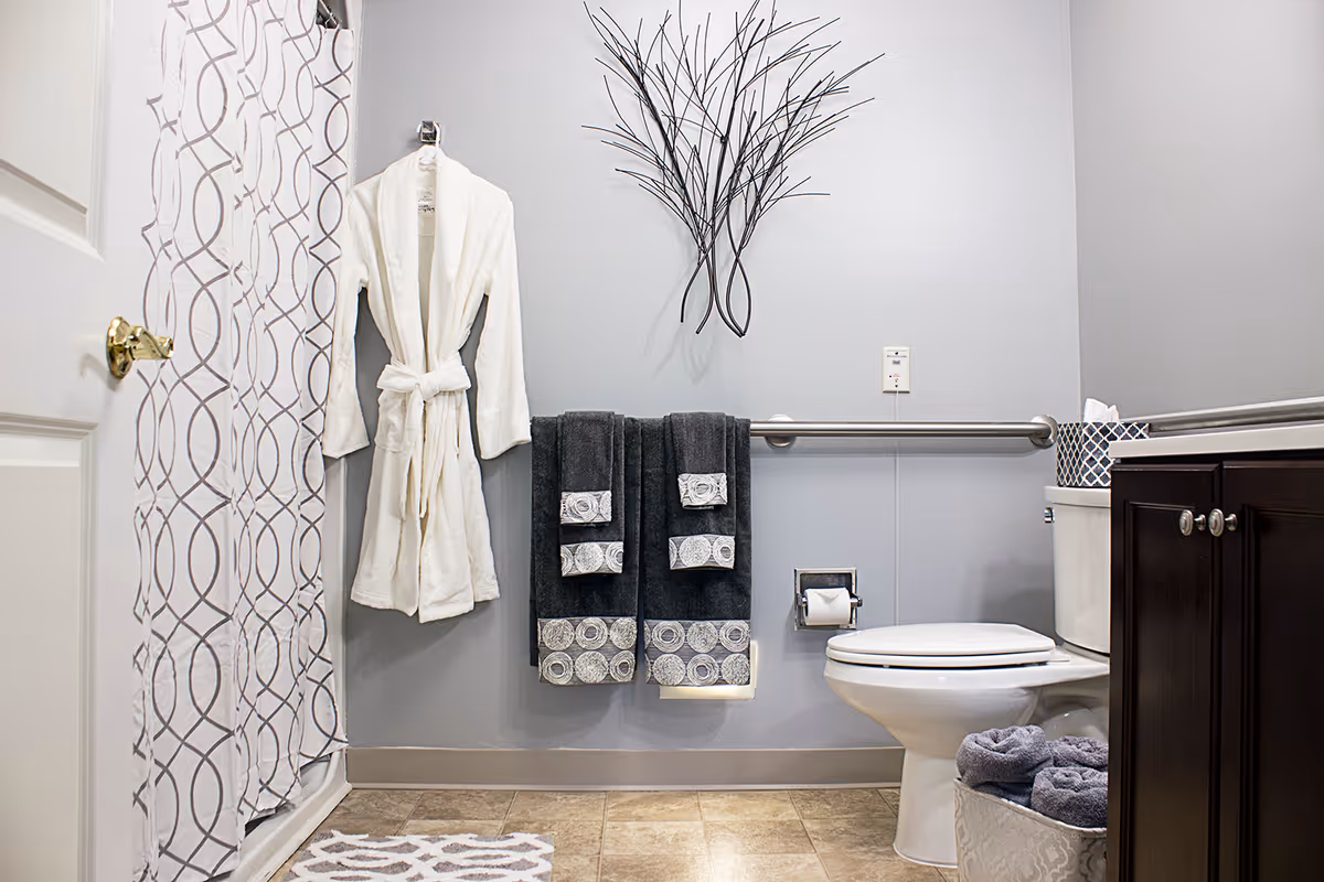 Bathroom with a patterned shower curtain, a hanging white robe, dark decorative towels, wall art, grab bar, and a toilet next to a dark vanity.