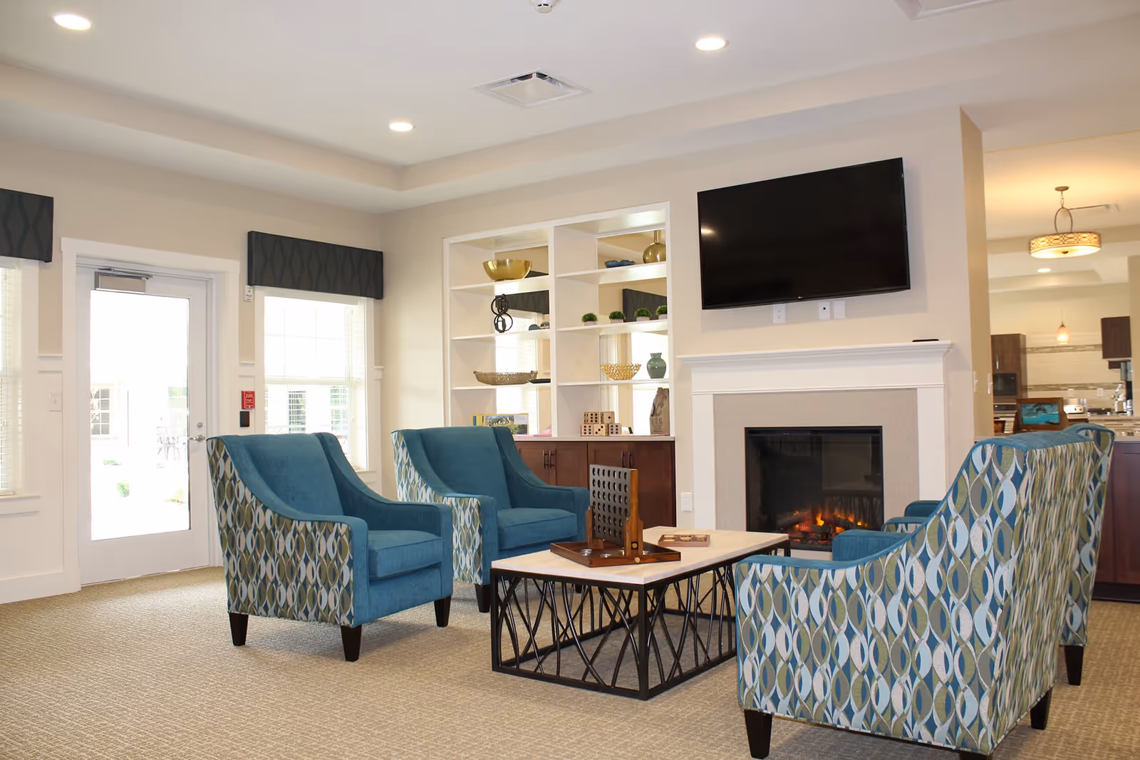 A cozy living room area with four blue patterned armchairs arranged around a rectangular coffee table with a game on it. There is a fireplace with a TV mounted above it, built-in shelves with decorative items, and large windows letting in natural light.