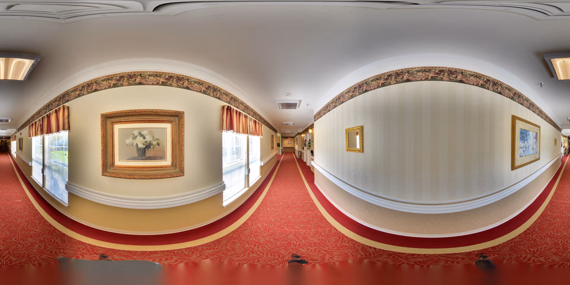 A long, carpeted hallway in a senior living facility with red patterned carpet and cream-colored walls. The walls are decorated with framed floral artwork and a floral wallpaper border near the ceiling. Windows with striped valances allow natural light into the hallway.
