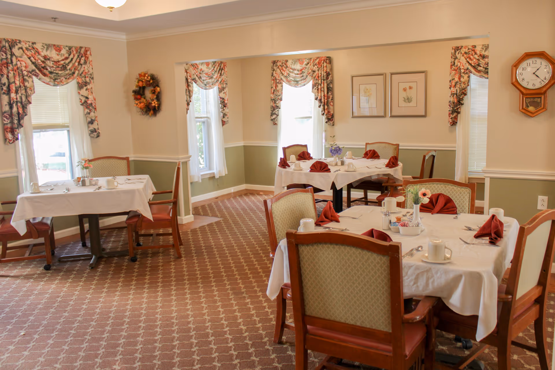 A dining room with several tables covered in white tablecloths, each set with cups, utensils, and red folded napkins. The room has patterned carpet, floral curtains on the windows, framed artwork on the walls, and a wooden clock hanging on the wall.