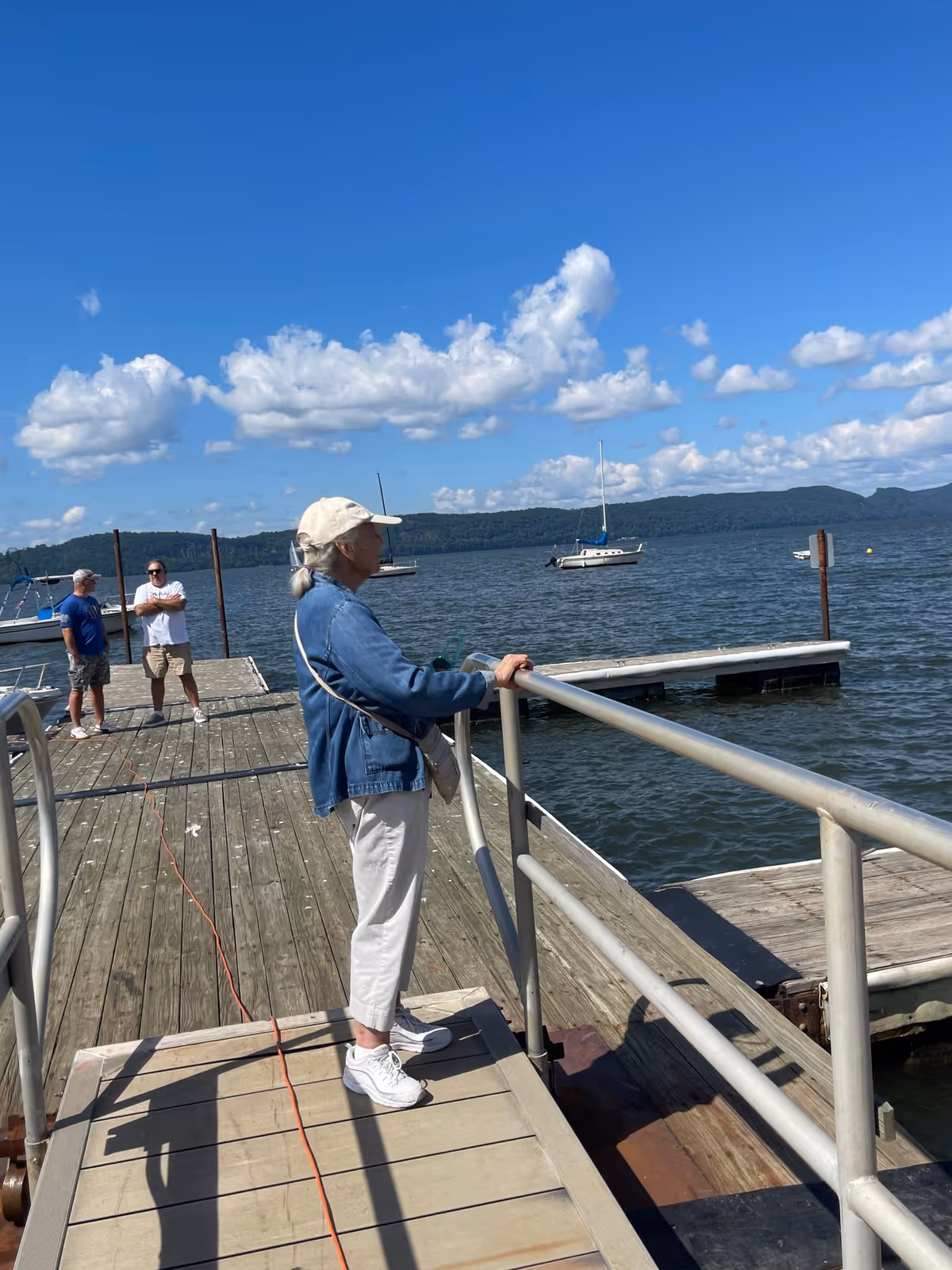 A woman wearing a white cap and denim jacket stands on a wooden dock looking out over a lake with sailboats under a blue sky.