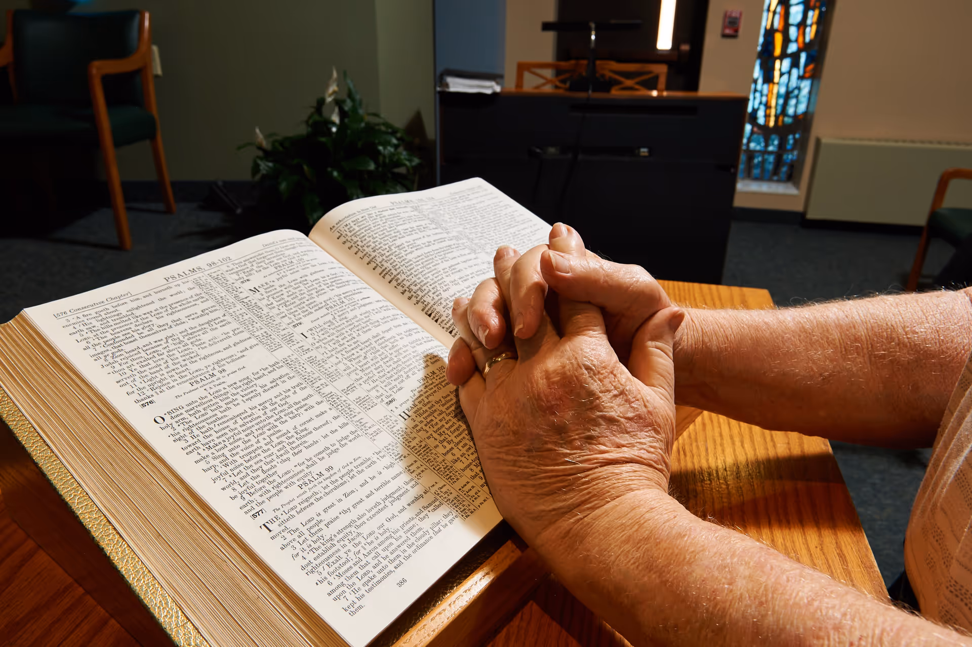 Close-up of an elderly person's hands clasped in prayer over an open Bible on a wooden lectern in a room with chairs, a plant, and a stained glass window in the background.