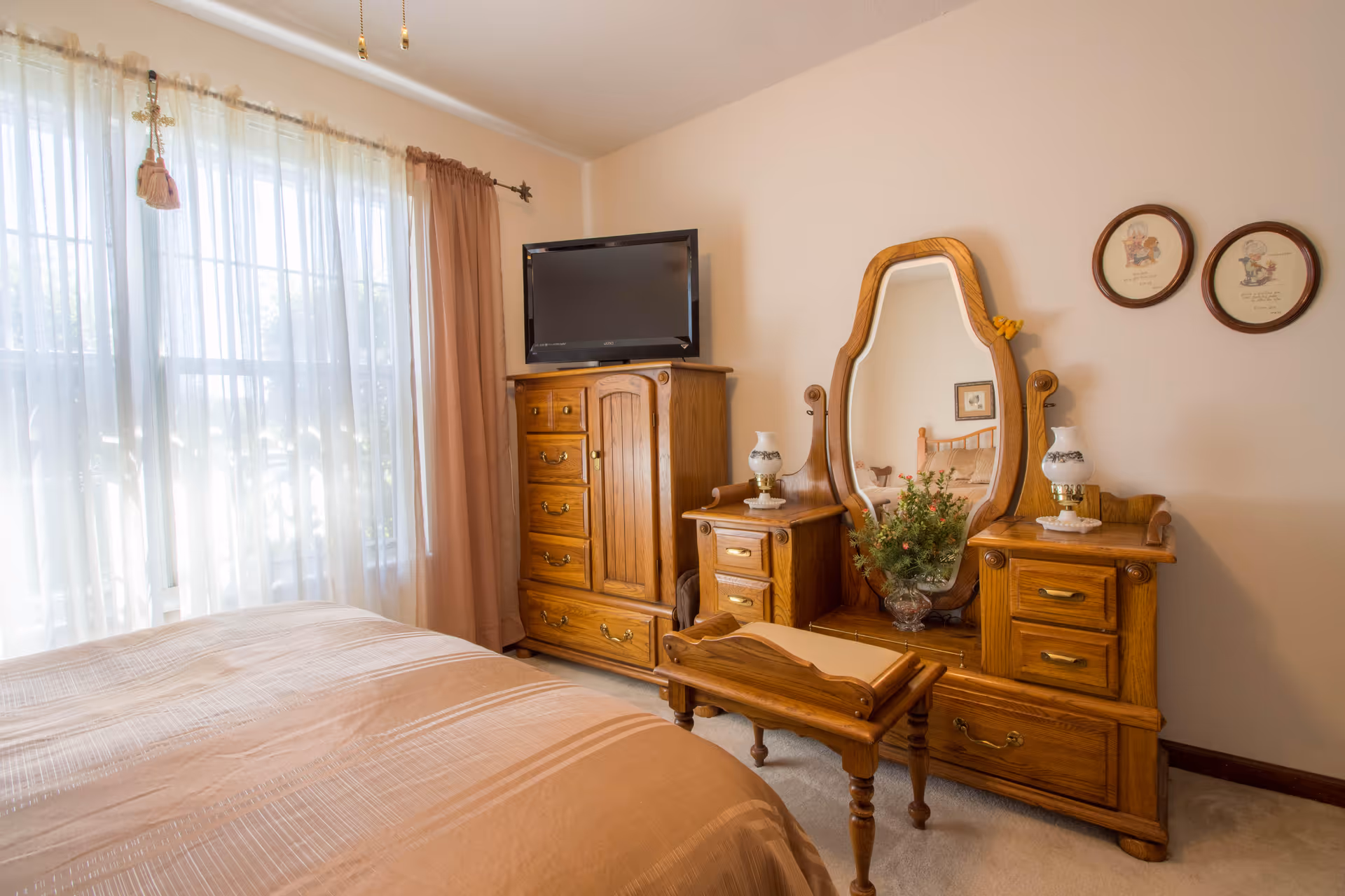 Sunlit bedroom featuring a wooden dresser and vanity with mirror, a TV, and a bed by a large curtained window.