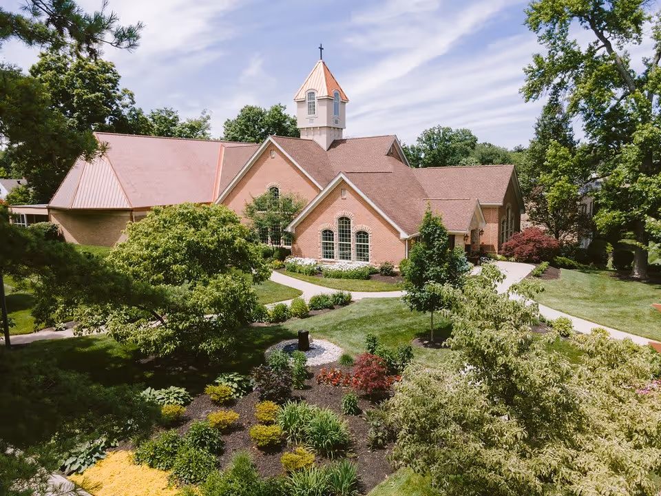 A brick building with a peaked roof and a small tower topped with a cross, surrounded by well-maintained gardens and trees under a partly cloudy sky.