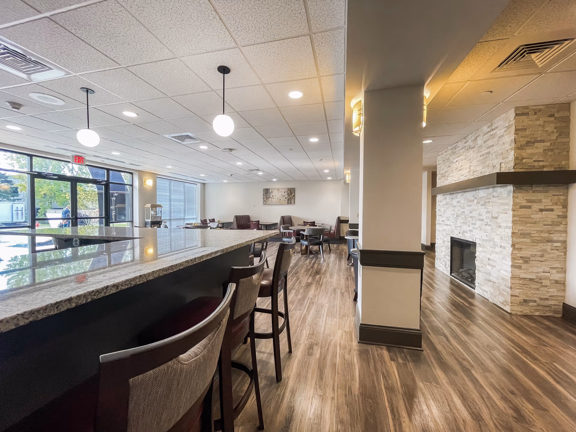 Interior view of a senior living facility common area featuring a long granite countertop with bar stools, a seating area with tables and chairs, large windows letting in natural light, and a stone fireplace on the right side.