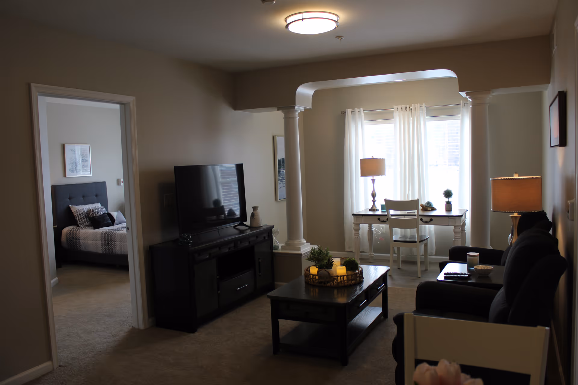 A cozy living room with a dark sofa and armchair, a coffee table with decorative candles and plants, a TV on a stand, and a small dining area near a window with white curtains. A bedroom with a bed and framed artwork is visible through an open doorway.