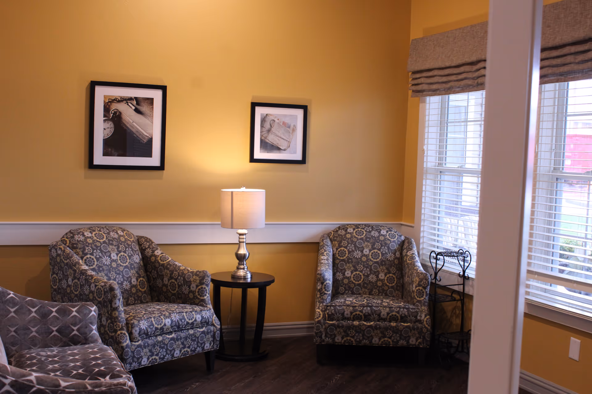 A cozy sitting area with two patterned armchairs and a small round table with a lamp between them. The walls are painted yellow and decorated with two framed pictures. There are windows with blinds on the right side letting in natural light.