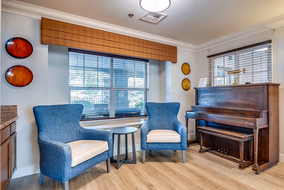 A cozy sitting area with two blue armchairs and a small round table between them, positioned in front of a large window with blinds. To the right, there is a wooden upright piano with a matching bench. The walls are decorated with four circular wall hangings, two on each side of the window. The floor is light wood, and the room is well-lit with natural light.