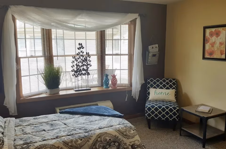 A cozy bedroom with a bed covered in a patterned comforter, a large window with blinds and sheer white curtains, a small potted plant and decorative items on the windowsill, a patterned armchair with a pillow that says 'home', a small side table with a book, and a framed floral picture on the wall.