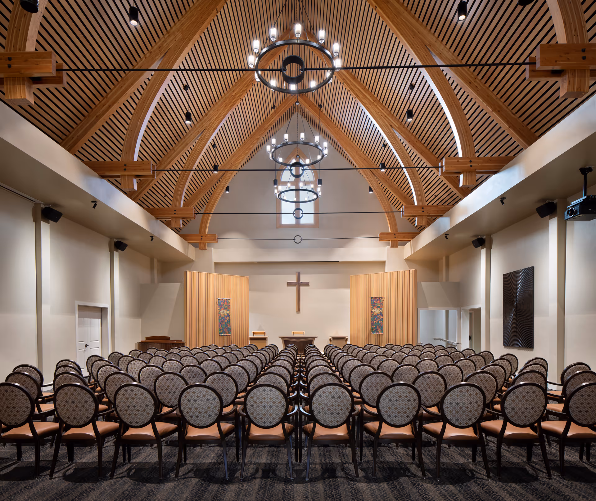 Interior chapel/auditorium with rows of upholstered chairs facing a raised platform with a cross under a vaulted wooden ceiling and chandeliers.