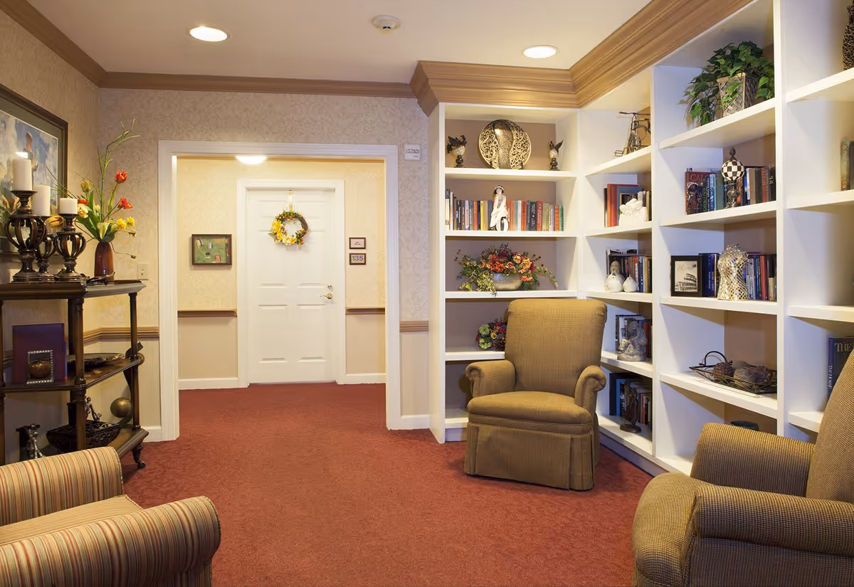 A cozy sitting area in a senior living facility with two upholstered armchairs facing each other, a built-in white bookshelf filled with books and decorative items, a small table with candles and flowers, and a white door decorated with a wreath in the background.