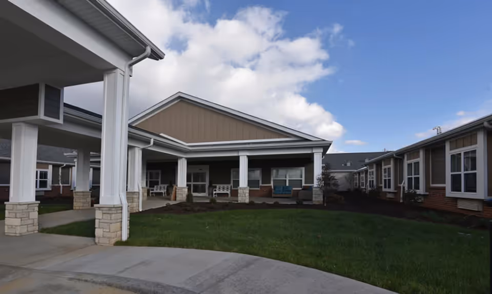 Exterior view of a senior living facility with a covered entrance supported by white columns with stone bases, a green lawn in front, and multiple windows on the building under a partly cloudy blue sky.