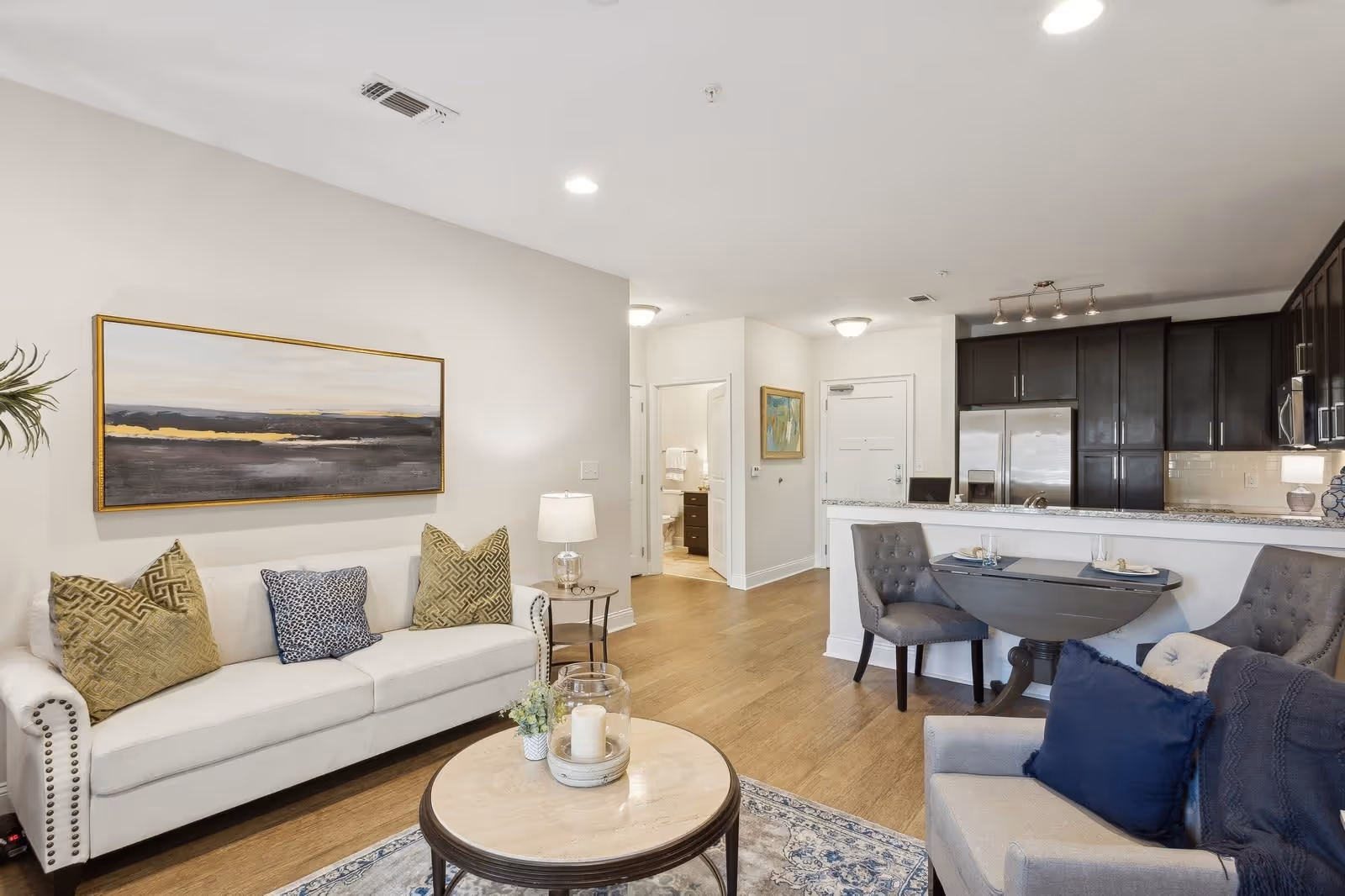 A modern living room and kitchen area in a senior living facility. The living room features a white sofa with decorative pillows, a round coffee table with candles and a small plant, and a gray armchair with blue cushions. The kitchen has dark wood cabinets, stainless steel appliances, and a granite countertop with two gray upholstered chairs at a small dining table. The walls are light-colored with a large abstract painting above the sofa and a framed picture near the entrance. The floor is wood, and the space is well-lit with ceiling lights.