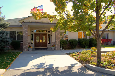 Front entrance of a single-story building with double wooden doors featuring oval glass panels, flanked by brick columns. There is an American flag flying above the entrance, surrounded by green bushes, a tree with green leaves, and a small garden area with flowers. The building appears to be part of a senior living facility named Edgewood Castle Hills.