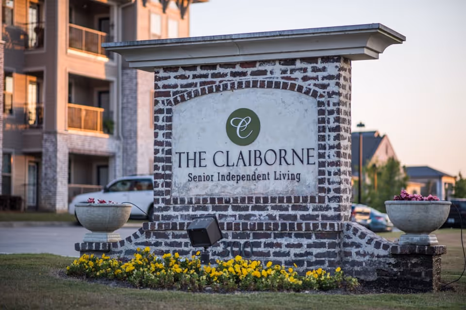 A brick sign for The Claiborne Senior Independent Living with flower beds and planters on either side, set in front of a multi-story residential building and parked cars.