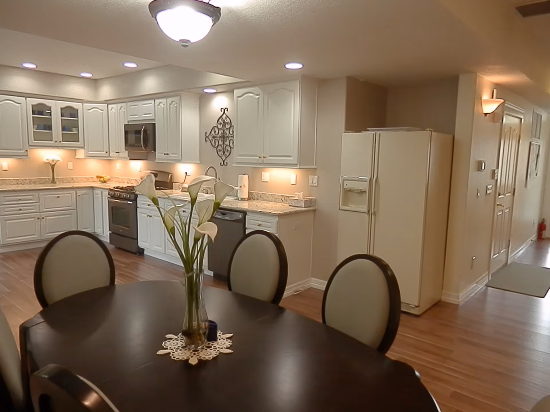 A modern kitchen and dining area with white cabinets, stainless steel appliances, a refrigerator, and a dark wooden dining table with four chairs. A vase with white flowers is placed on the dining table. The room has wooden flooring and recessed lighting.