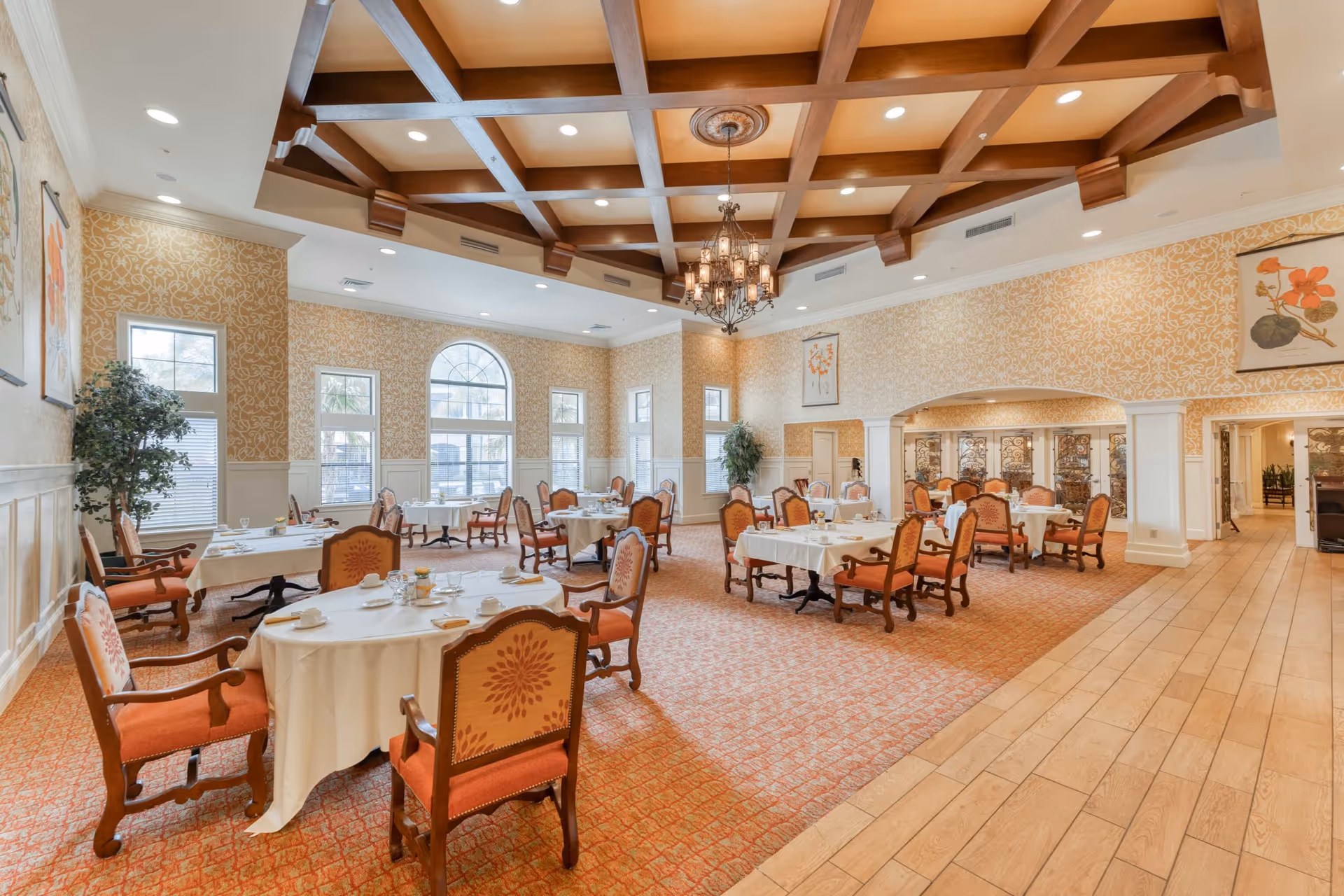 Spacious elegant dining room with round tables set with white tablecloths, upholstered chairs, arched windows and a coffered ceiling chandelier.