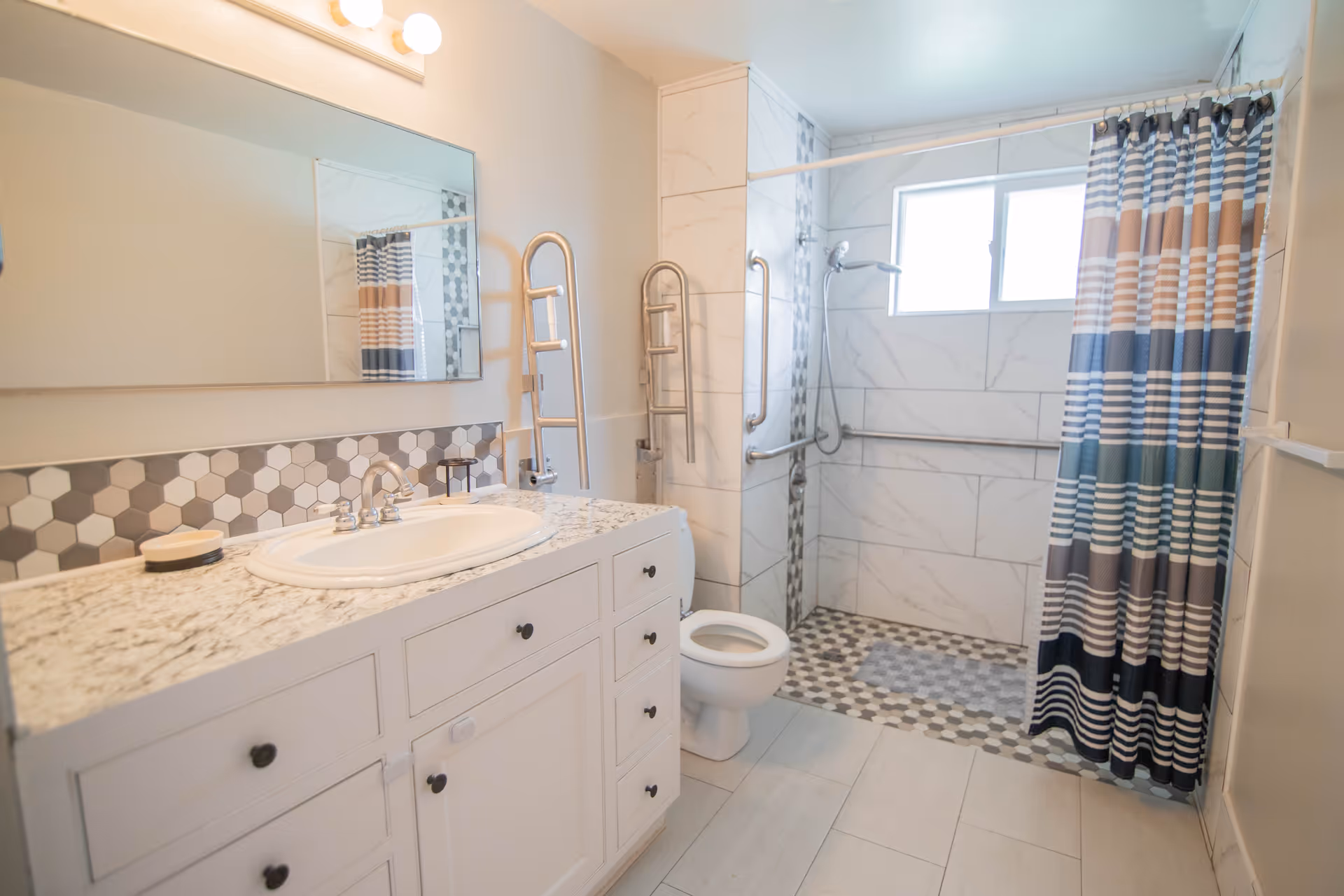 Accessible bathroom with a white vanity and sink, a toilet, grab bars and a tiled walk-in shower with a striped shower curtain.