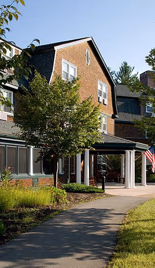 Exterior view of a multi-story building with brown shingles and white-trimmed windows, surrounded by trees and greenery. A paved walkway leads to a covered entrance supported by white columns. An American flag is visible on the right side near the entrance.