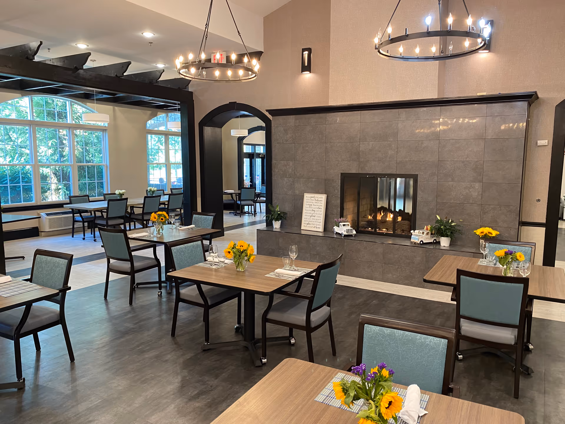 Bright dining room with multiple set tables and chairs, a central fireplace, chandeliers, and large sunlit windows.
