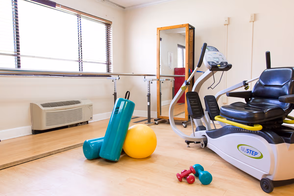 Bright exercise room with a recumbent NuStep machine, stability balls, dumbbells, mirrors and handrails.