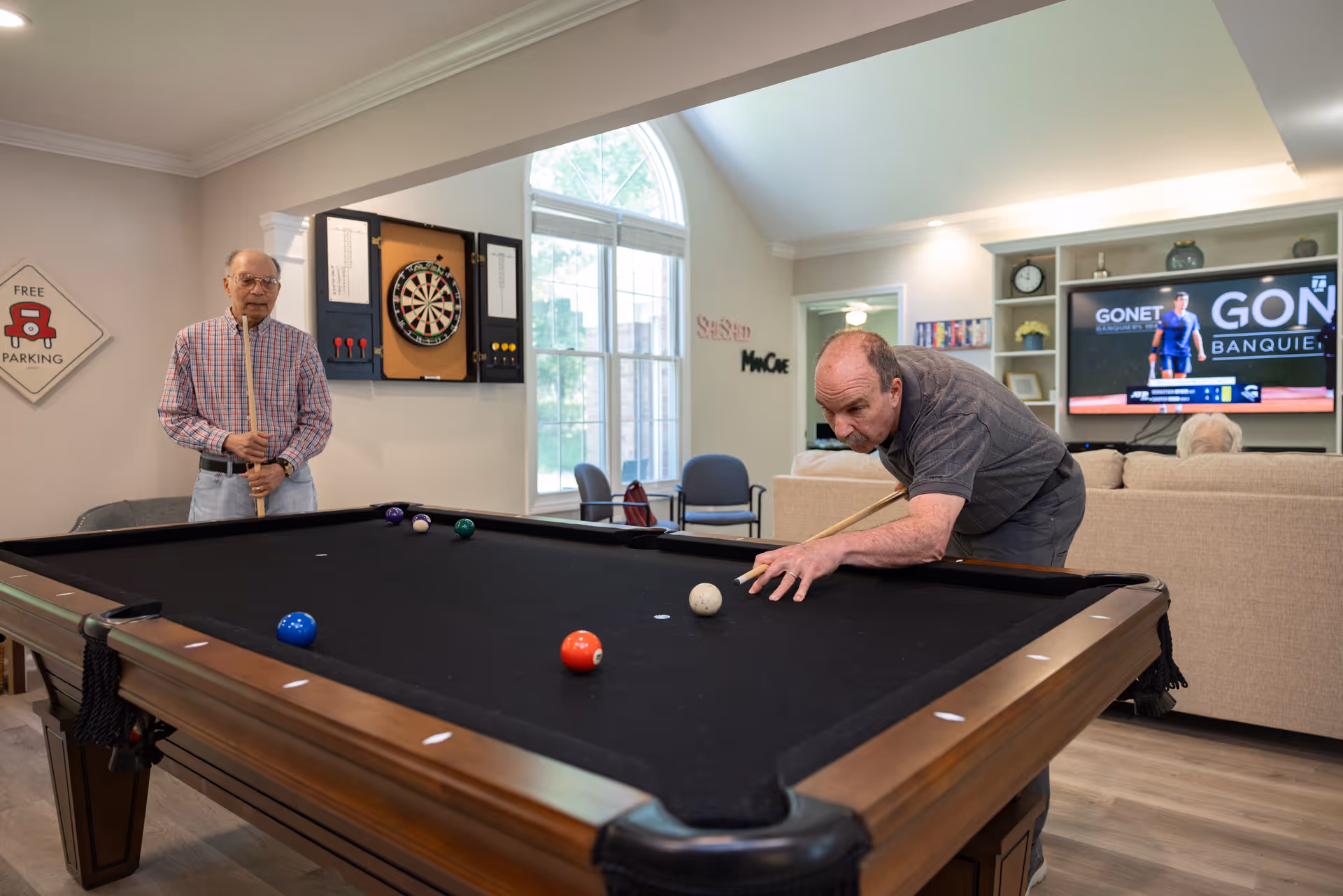 Two elderly men playing pool in a well-lit recreational room with a dartboard on the wall, a large window, and a television in the background showing a sports game. One man is preparing to take a shot while the other watches. The room has comfortable seating and a sign on the wall that says 'Free Parking.'