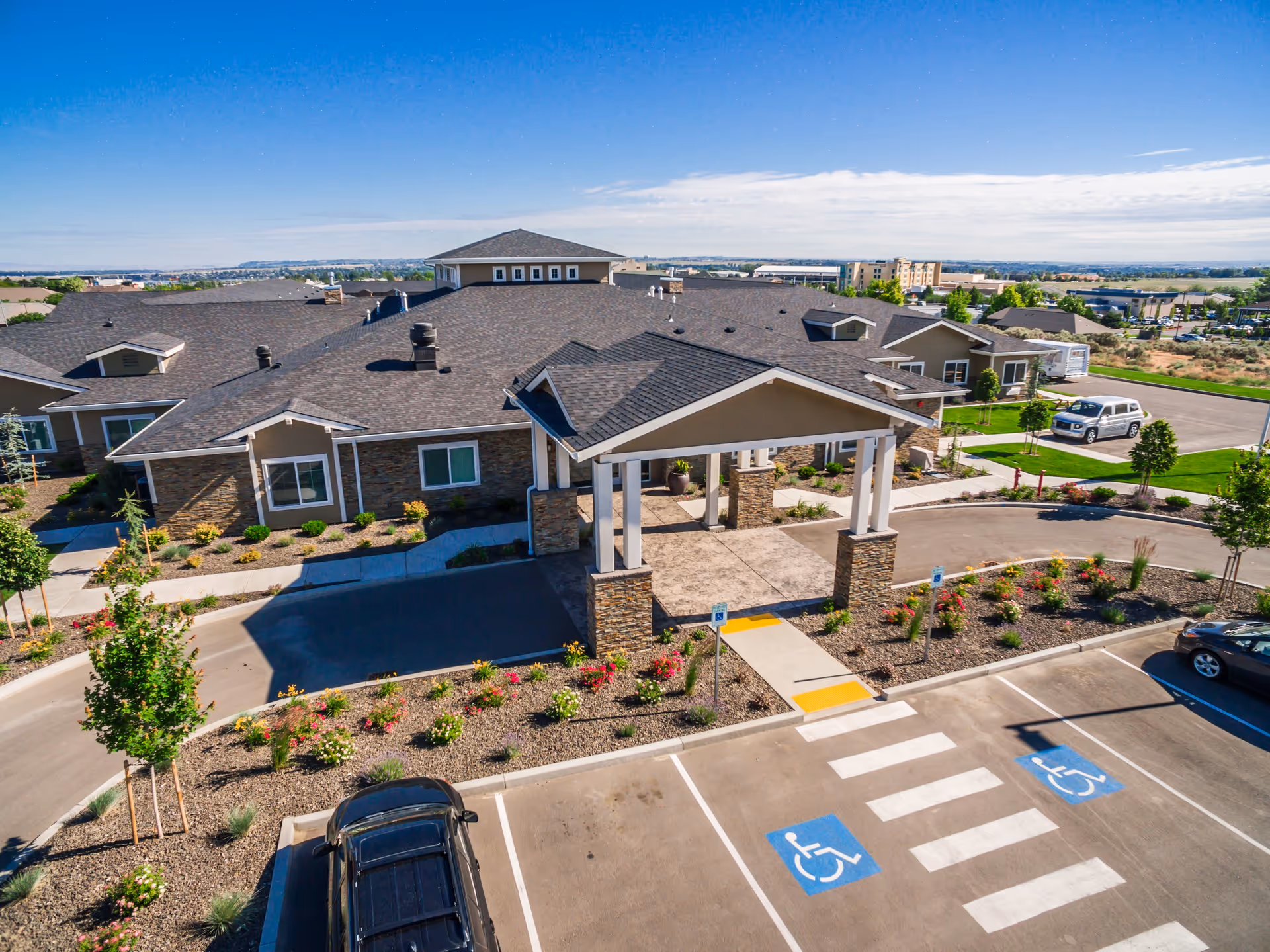 Aerial view of the entrance to Ciel of Tri-Cities Memory Care facility showing a covered drop-off area with stone pillars, landscaped garden beds with flowers and shrubs, a parking lot with handicap parking spaces, and surrounding buildings under a clear blue sky.