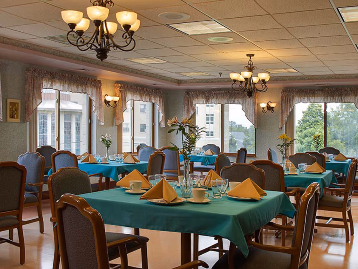 A dining room with multiple tables covered in teal tablecloths, each set with folded yellow napkins, cups, glasses, and small flower vases. The room has large windows with floral curtains, wooden chairs with cushioned seats, and chandeliers hanging from the ceiling providing warm lighting.