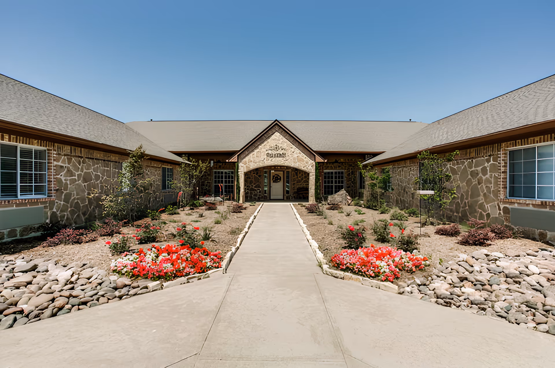 Front entrance of a single-story stone building with a central concrete walkway and landscaped flower beds under a clear blue sky.