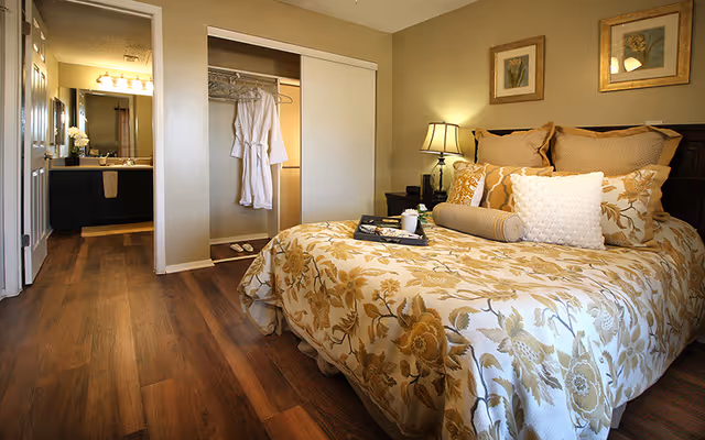 Well-lit bedroom with a floral bedspread, bedside lamp, open closet with a hanging robe, and a view into the bathroom.
