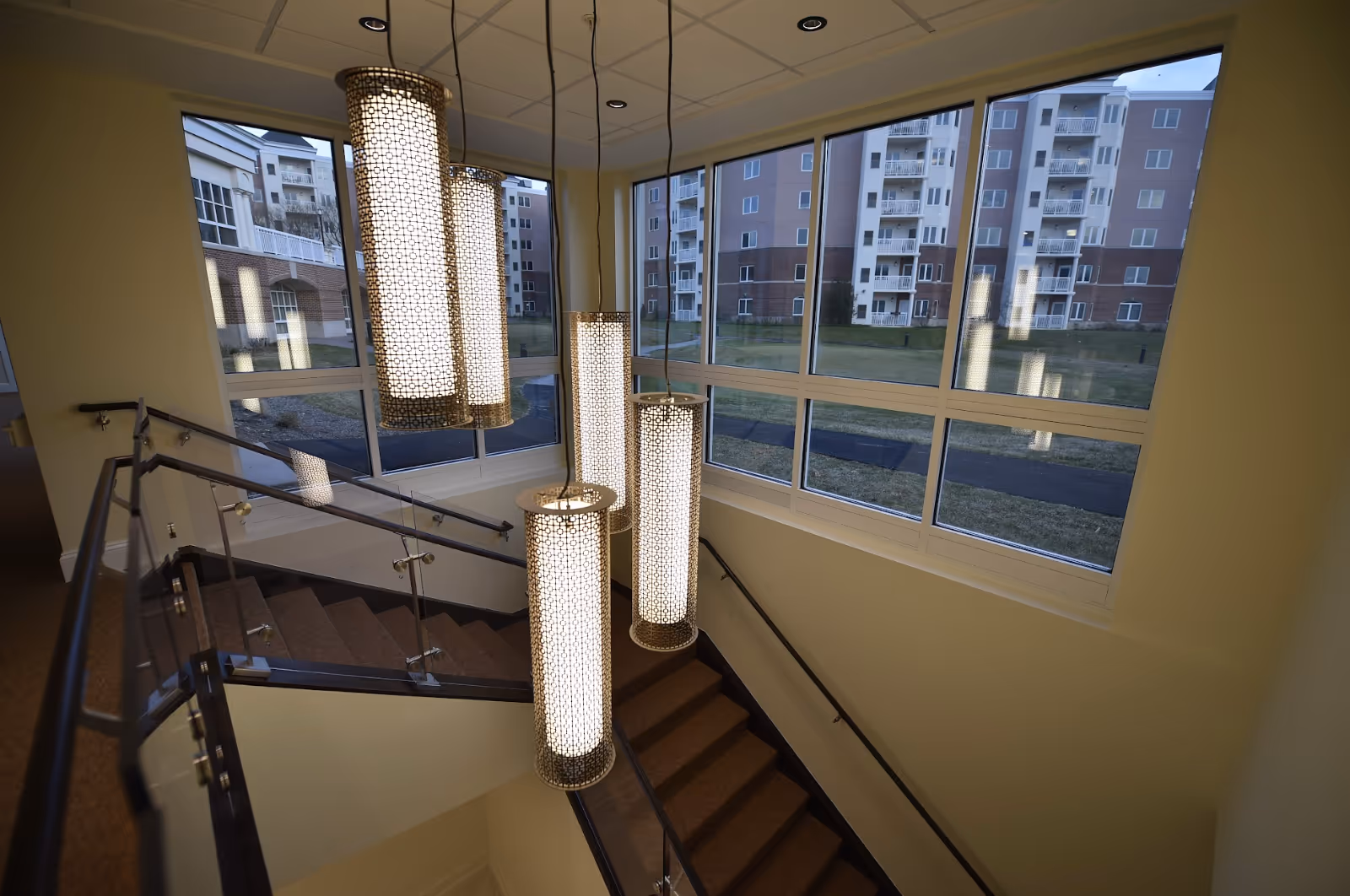 Interior stairwell with hanging cylindrical chandeliers and large windows overlooking neighboring apartment buildings.