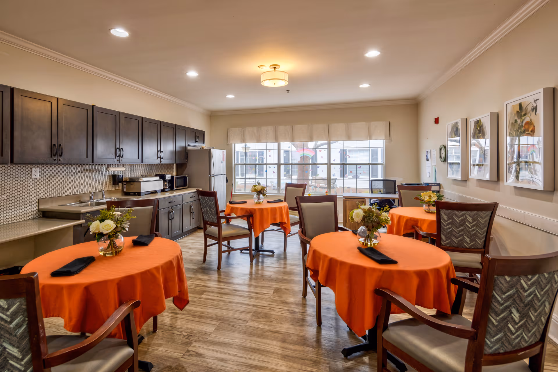 A bright dining room with four round tables covered in orange tablecloths, each set with a small floral centerpiece and a black napkin. The room features wooden chairs with cushioned seats, a large window with a beige valance letting in natural light, and a kitchen area along one wall with dark wooden cabinets, a refrigerator, microwave, and countertop appliances. The walls are decorated with framed artwork.