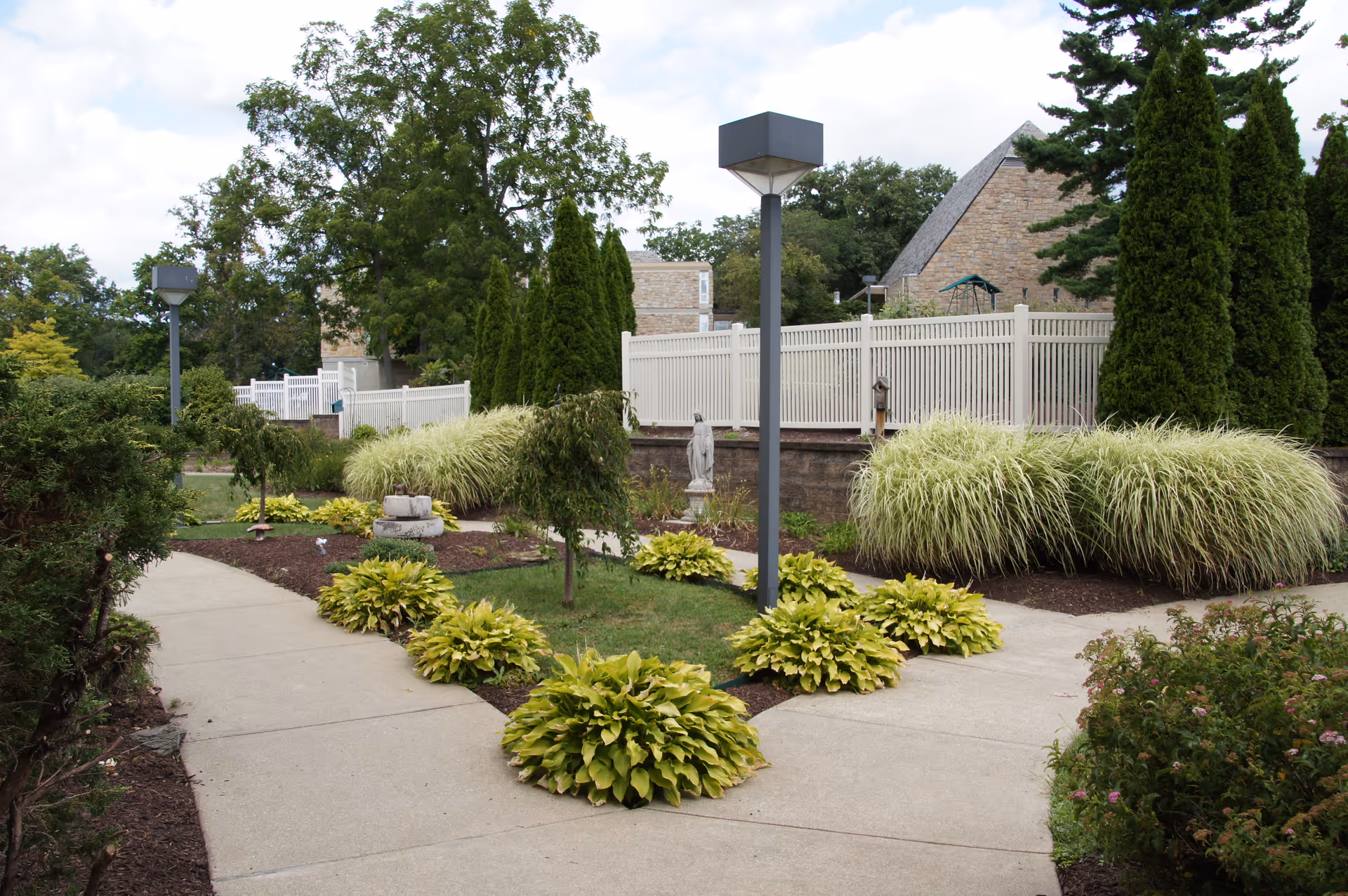 Outdoor garden area at Elizabeth Seton Memory Care Center featuring paved walkways, green shrubs, ornamental grasses, a small statue, and tall trees with a white fence and stone buildings in the background under a partly cloudy sky.