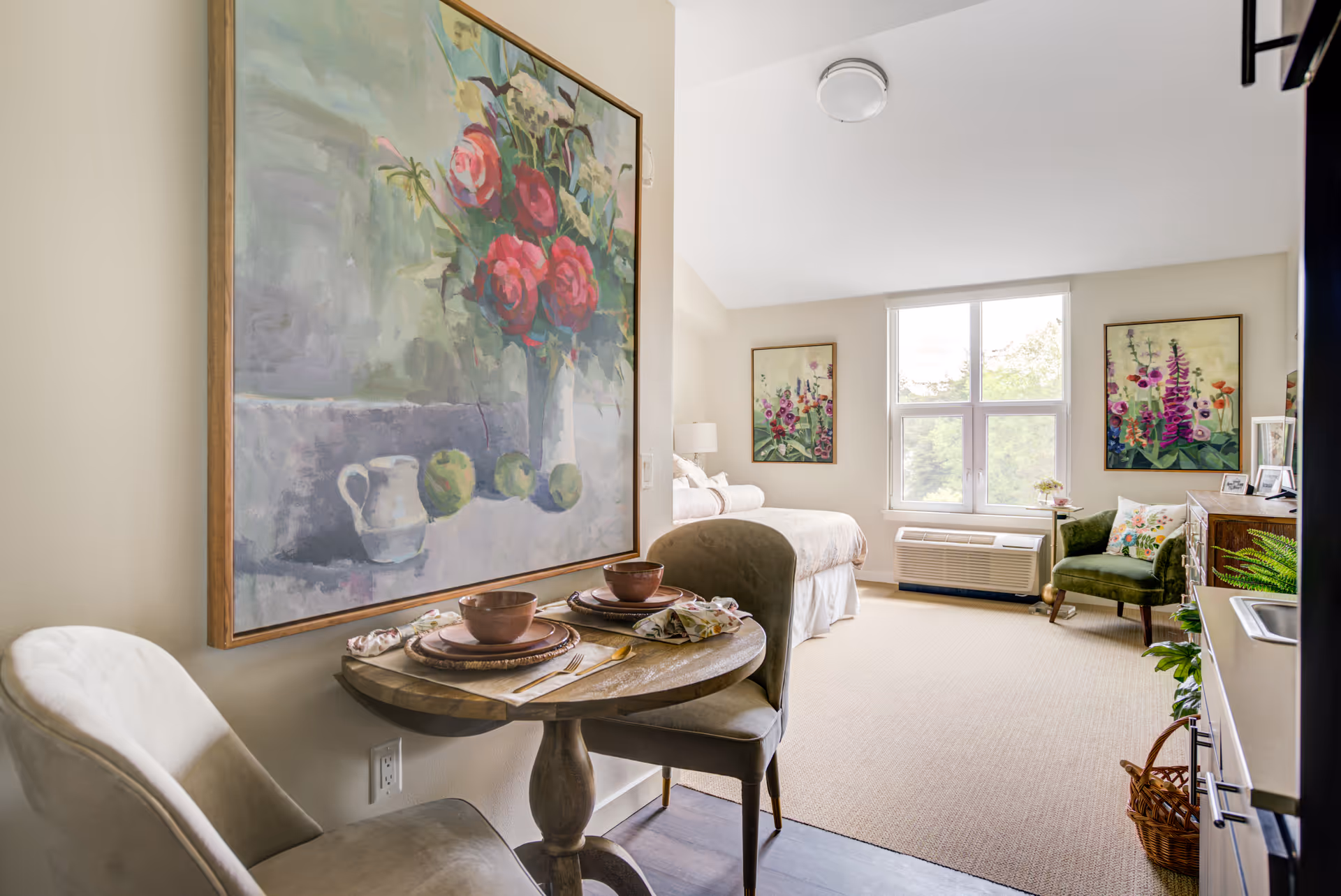 A bright and cozy studio apartment featuring a small round dining table set for two with plates, bowls, and napkins. The room includes a bed near a large window with natural light, a green armchair, a wooden dresser, and colorful floral paintings on the walls. A compact kitchenette with a sink and cabinets is visible on the right side.
