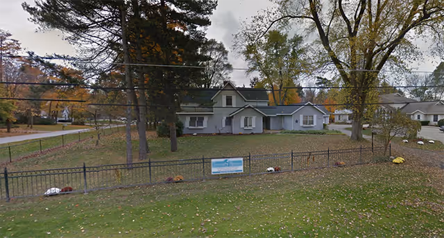 A single-story blue-gray senior living home behind a black metal fence with trees.